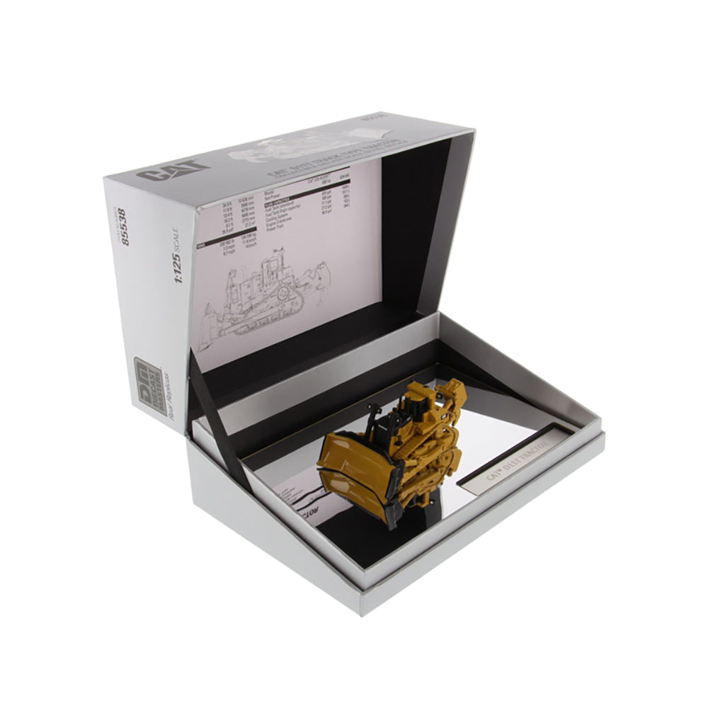 A miniature yellow excavator model displayed inside an open white presentation box with printed inserts and a mirror base, photographed on a clean white background.