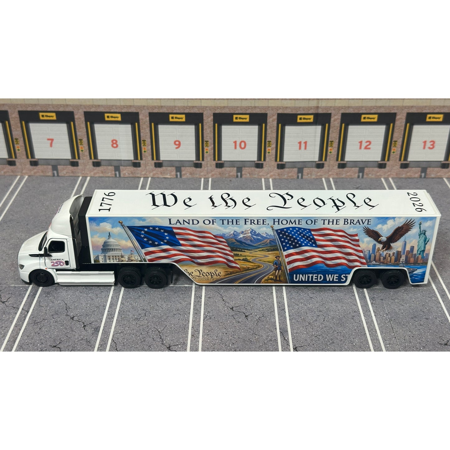 A diecast model of a Freightliner Cascadia semi-truck parked in a simulated parking lot. The trailer features a detailed historical design celebrating the United States' 250th anniversary with 'We the People' written on the top, alongside an American flag, eagle, and city skyline graphic with the text 'Land of the Free Home of the Brave' and 'United We Stand'.