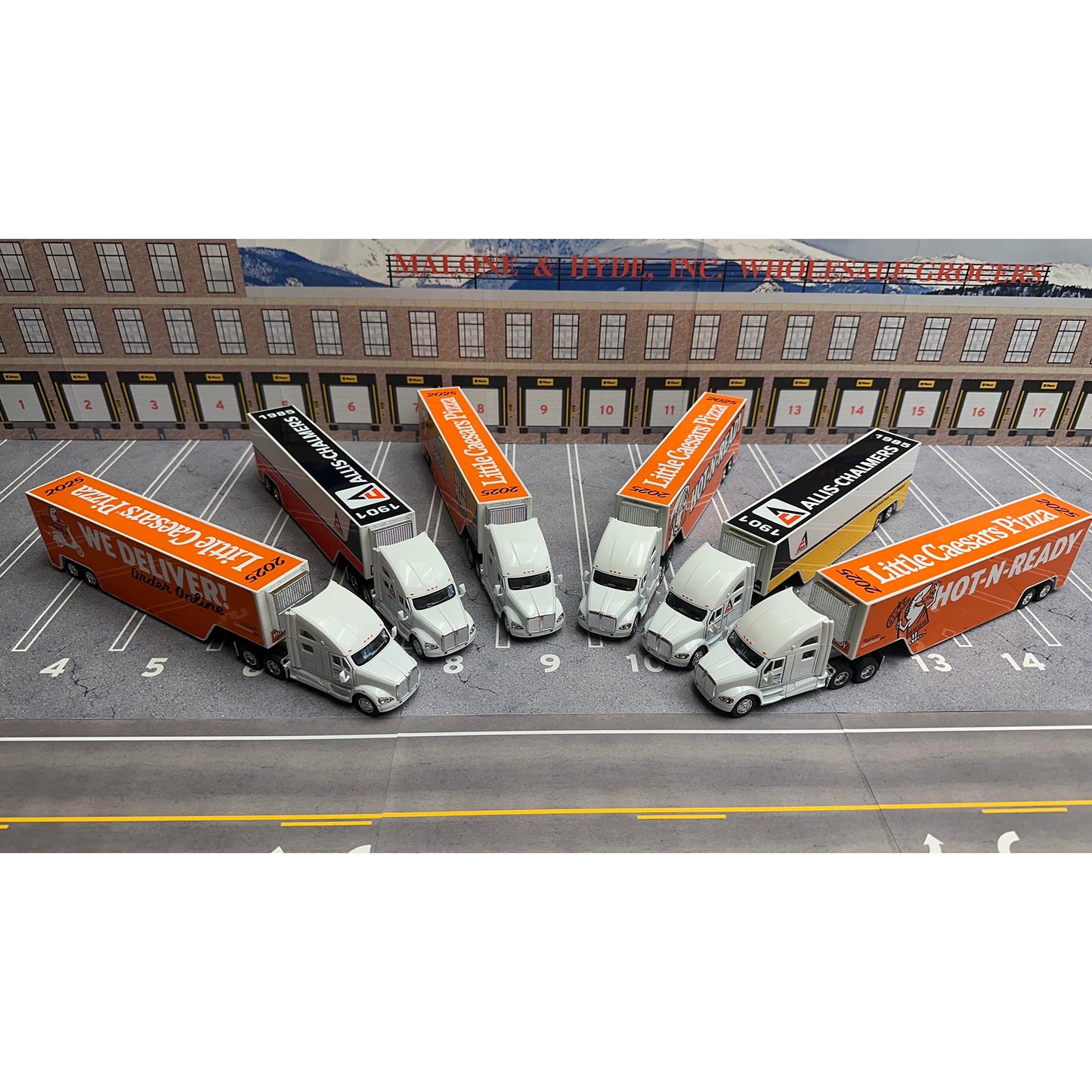 Model trucks with orange and white trailers on a parking lot with a building in the background