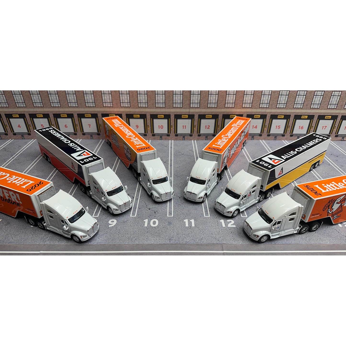 Model trucks and cars on a parking lot scale model with buildings in the background.