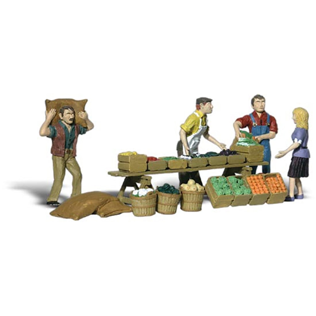 A miniature model market scene with four small figurines standing around a produce stand and bales of goods, photographed on a plain white background.