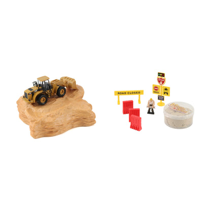 A miniature construction-themed toy set on a white background, featuring a yellow and black wheel loader on a tan rock base, along with road closure signs, red barriers, a small worker figure, and a round container of sand.