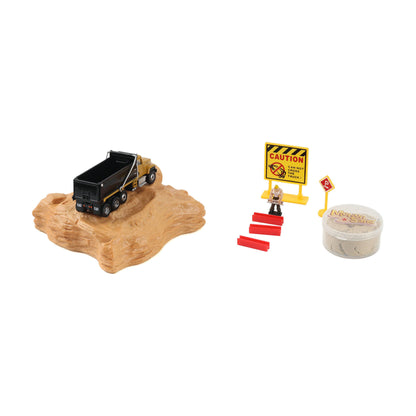 A toy construction scene set on a white background, featuring a black dump truck on a tan rock base, a yellow caution sign with a small worker figure, red steps, and a clear plastic container of toy bricks.