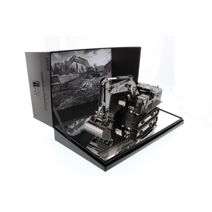 A  model of a black and silver excavator displayed inside its open black presentation box, which features a black-and-white construction scene printed on the inner flap.