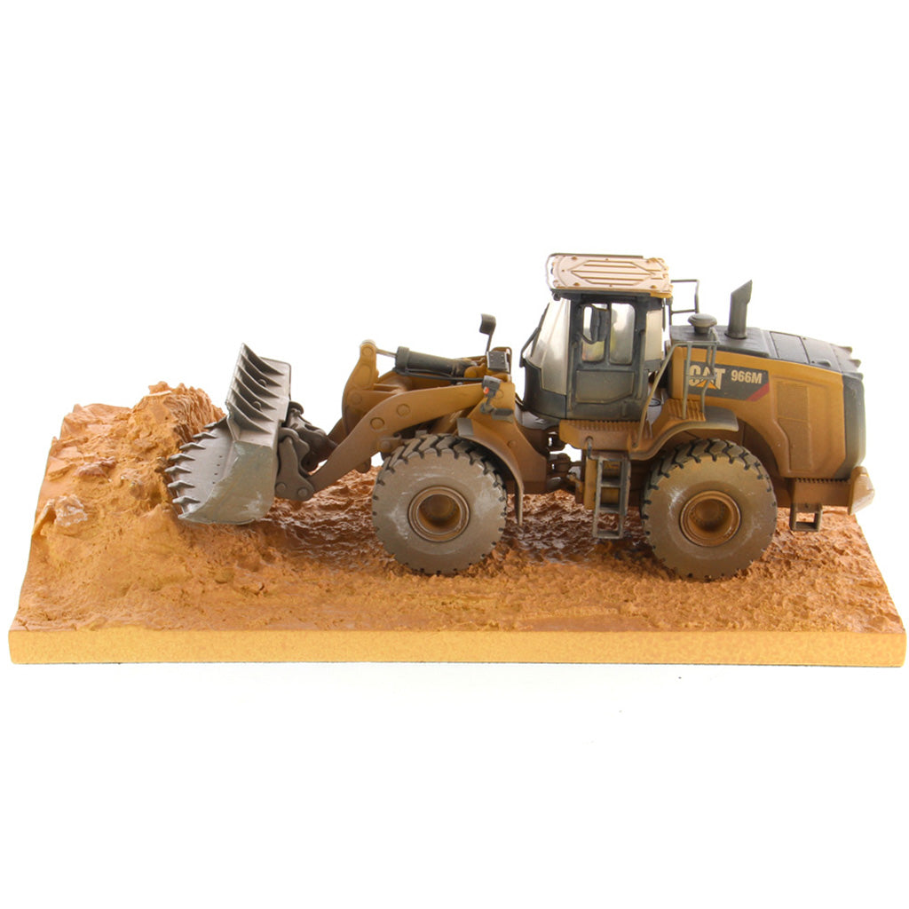 1:50th Scale Diecast Model of a Caterpillar 966M Wheel Loader with a Weathered Paint Scheme on a Tan Construction Site Base Plate on a White Background