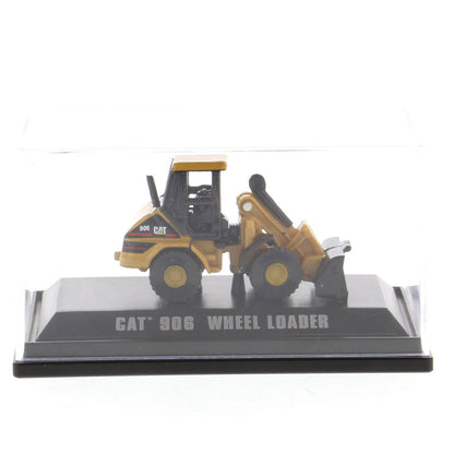 A miniature yellow and black CAT 906 wheel loader toy model displayed on a black base with 'CAT 906 WHEEL LOADER' printed on it, under a clear plastic cover.
