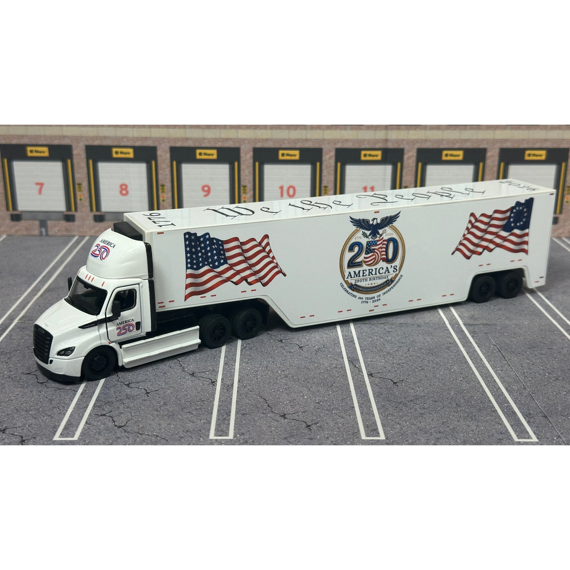 A diecast model of a white Freightliner Cascadia semi-truck with a long trailer decorated with American flags and a '250th Anniversary' logo, parked in a gray parking lot with a truck dock backdrop.