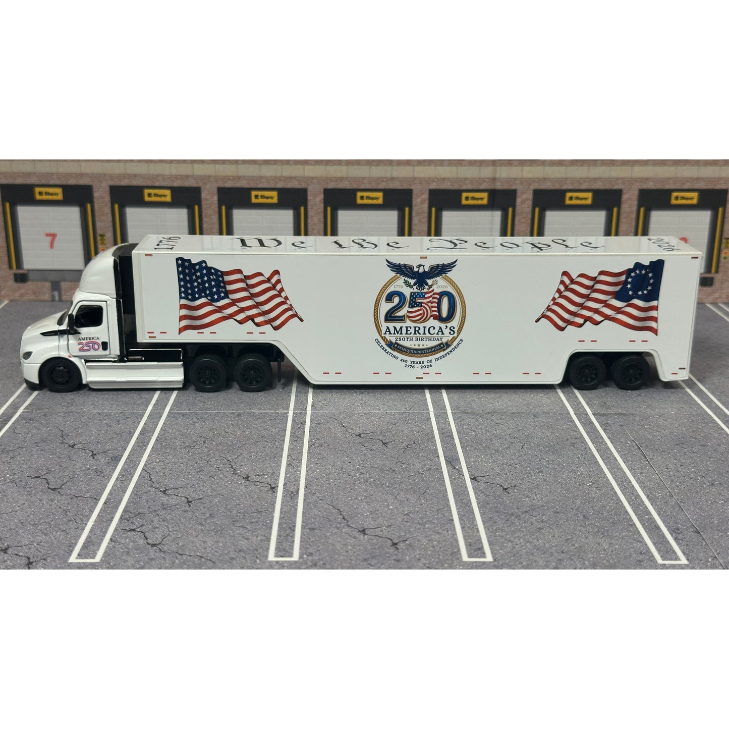 A diecast model of a white Freightliner Cascadia semi-truck with a long trailer decorated with American flags and a '250th Anniversary' logo, parked in a gray parking lot with a truck dock backdrop.