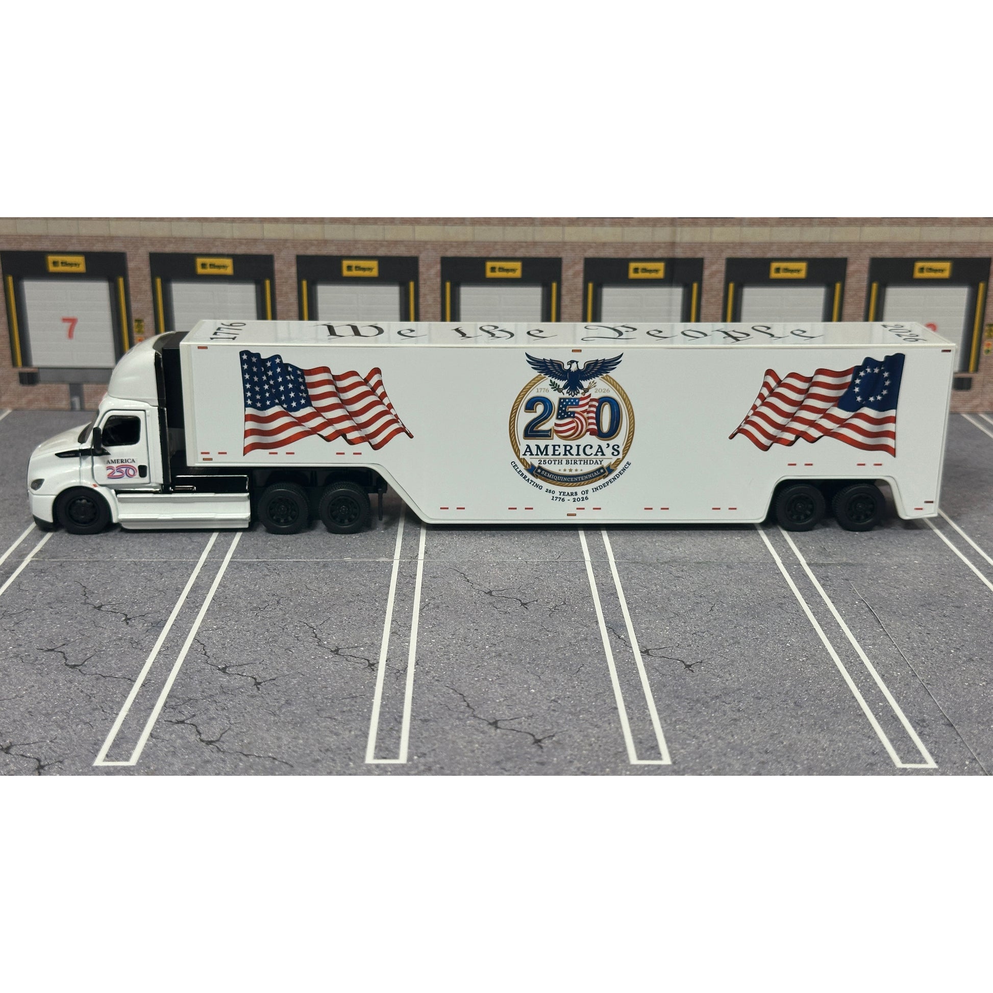 A diecast model of a white Freightliner Cascadia semi-truck with a long trailer decorated with American flags and a '250th Anniversary' logo, parked in a gray parking lot with a truck dock backdrop.