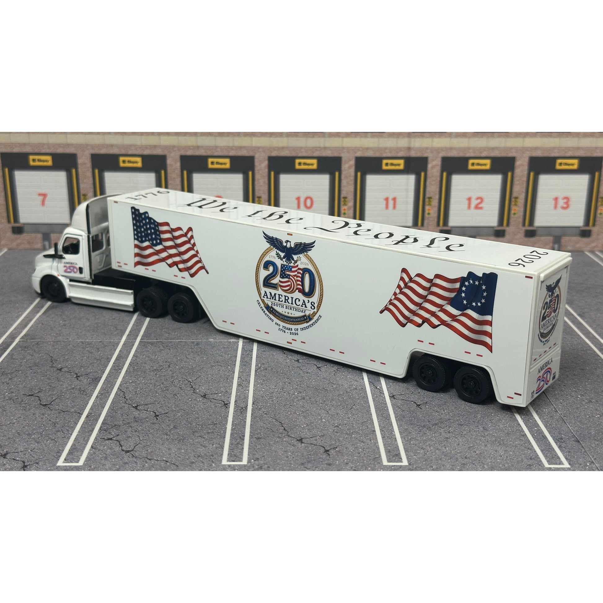 A diecast model of a white Freightliner Cascadia semi-truck with a long trailer decorated with American flags and a '250th Anniversary' logo, parked in a gray parking lot with a truck dock backdrop.