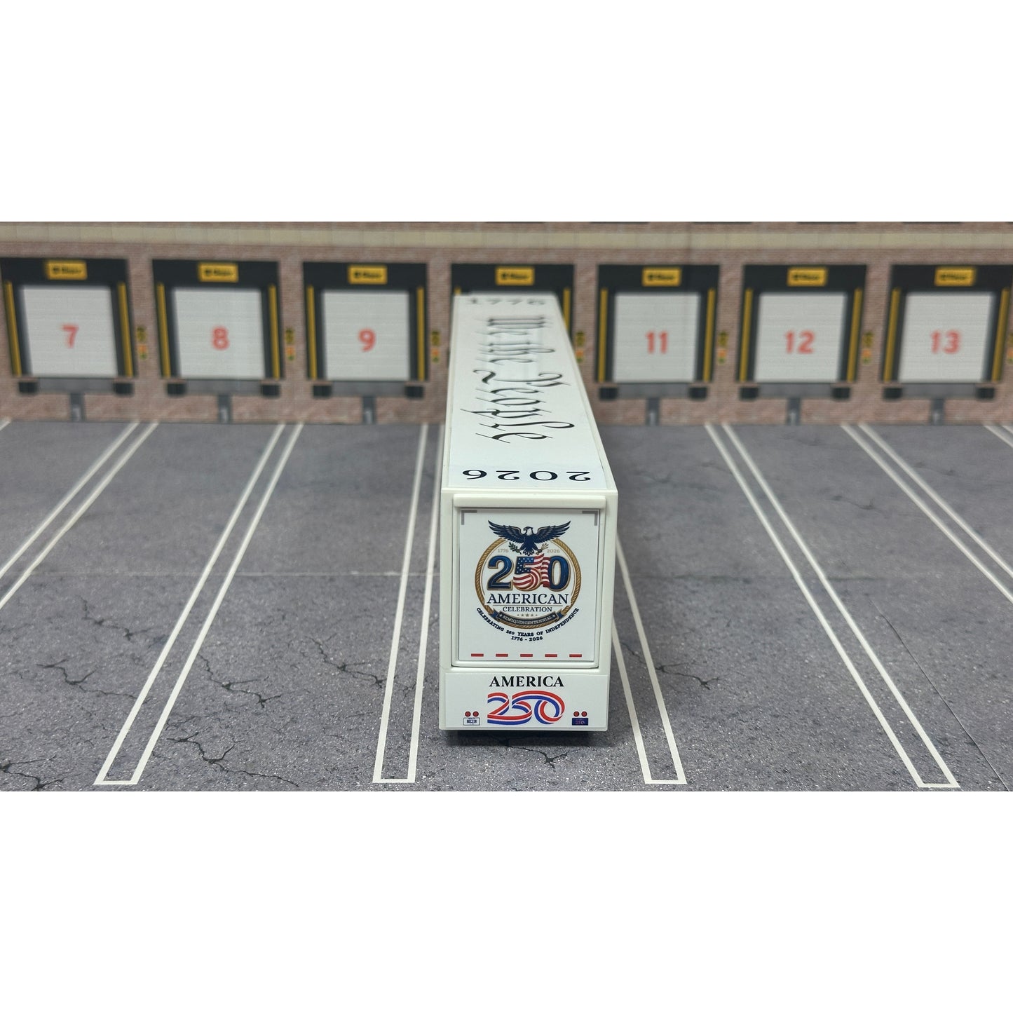A diecast model of a white Freightliner Cascadia semi-truck with a long trailer decorated with American flags and a '250th Anniversary' logo, parked in a gray parking lot with a truck dock backdrop.
