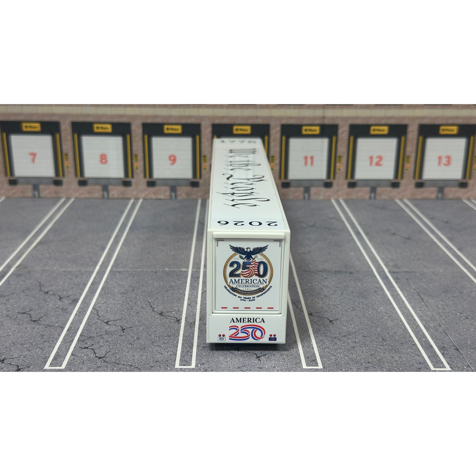 A diecast model of a white Freightliner Cascadia semi-truck with a long trailer decorated with American flags and a '250th Anniversary' logo, parked in a gray parking lot with a truck dock backdrop.