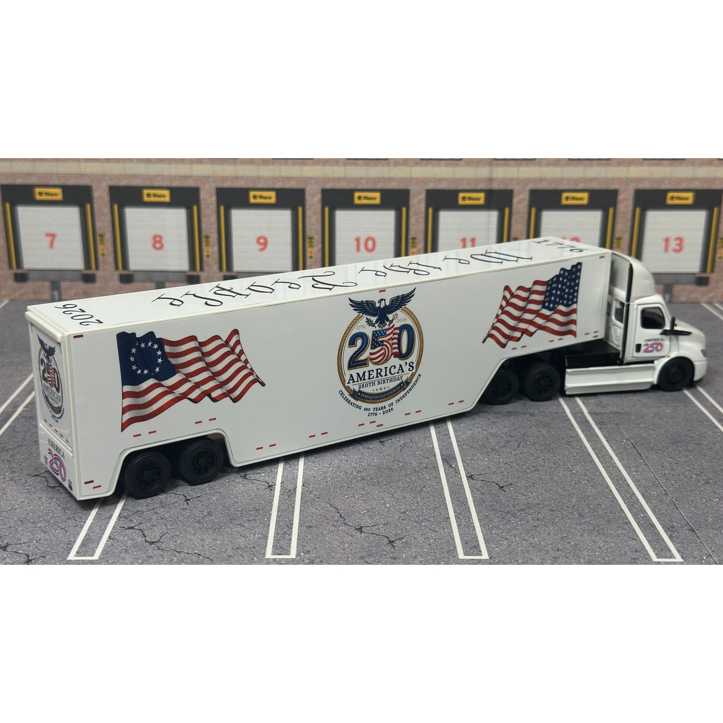 A diecast model of a white Freightliner Cascadia semi-truck with a long trailer decorated with American flags and a '250th Anniversary' logo, parked in a gray parking lot with a truck dock backdrop.