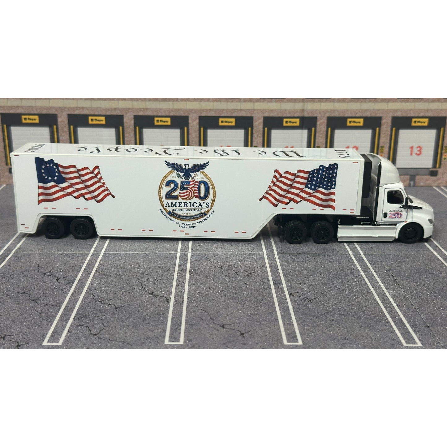 A diecast model of a white Freightliner Cascadia semi-truck with a long trailer decorated with American flags and a '250th Anniversary' logo, parked in a gray parking lot with a truck dock backdrop.