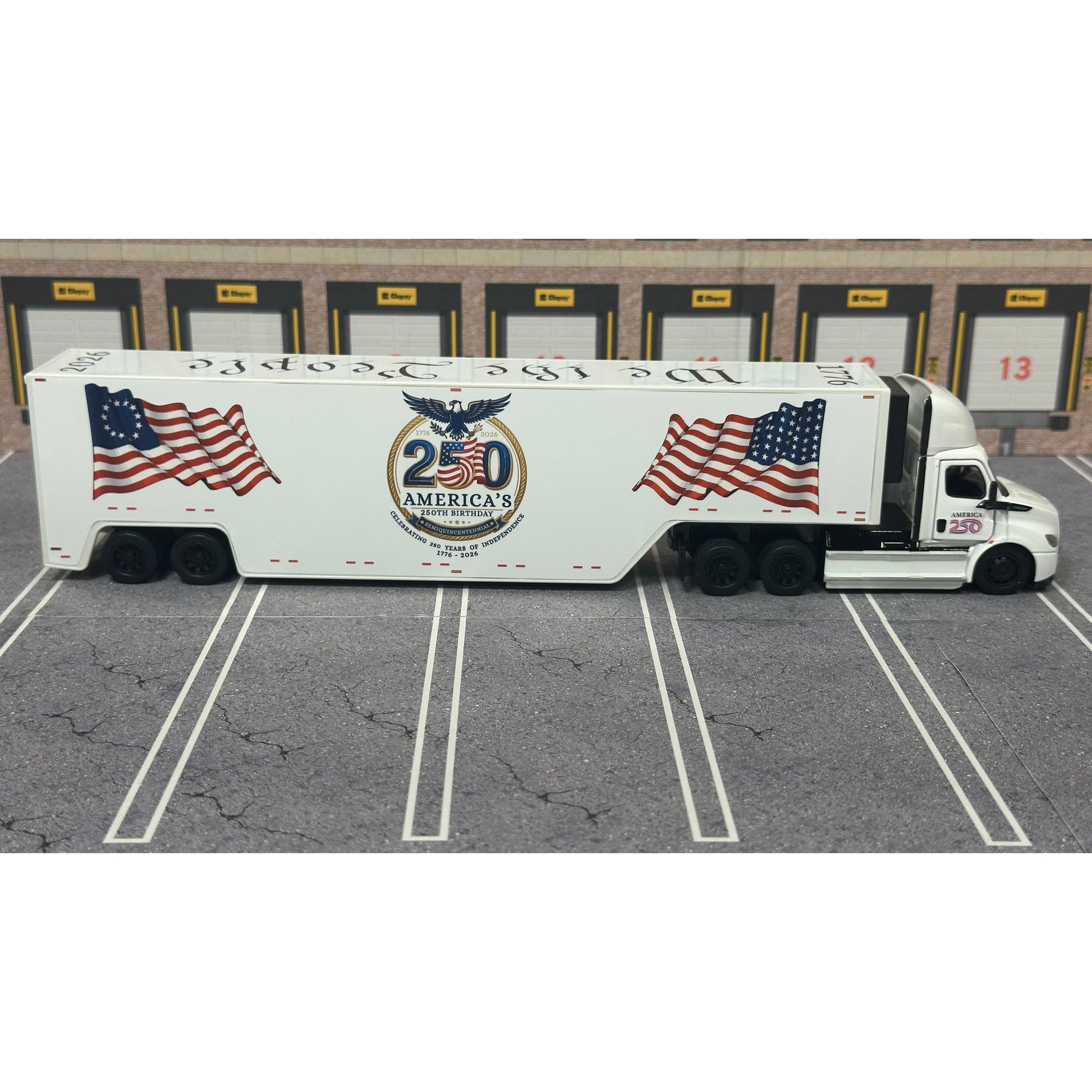 A diecast model of a white Freightliner Cascadia semi-truck with a long trailer decorated with American flags and a '250th Anniversary' logo, parked in a gray parking lot with a truck dock backdrop.
