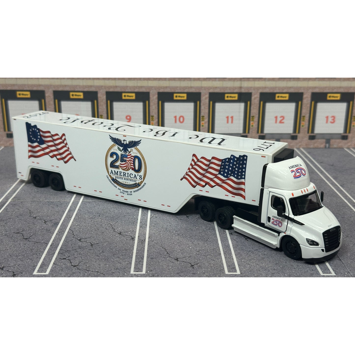 A diecast model of a white Freightliner Cascadia semi-truck with a long trailer decorated with American flags and a '250th Anniversary' logo, parked in a gray parking lot with a truck dock backdrop.