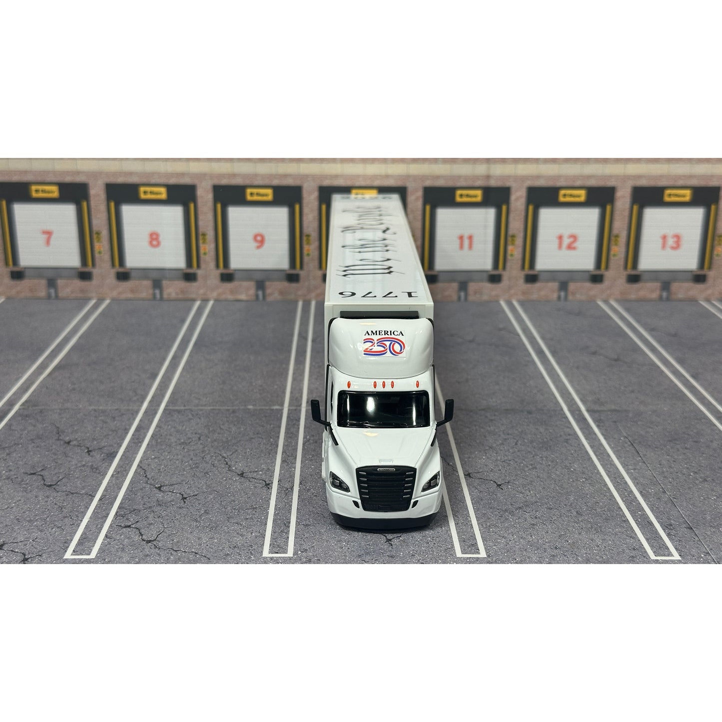 A diecast model of a white Freightliner Cascadia semi-truck with a long trailer decorated with American flags and a '250th Anniversary' logo, parked in a gray parking lot with a truck dock backdrop.