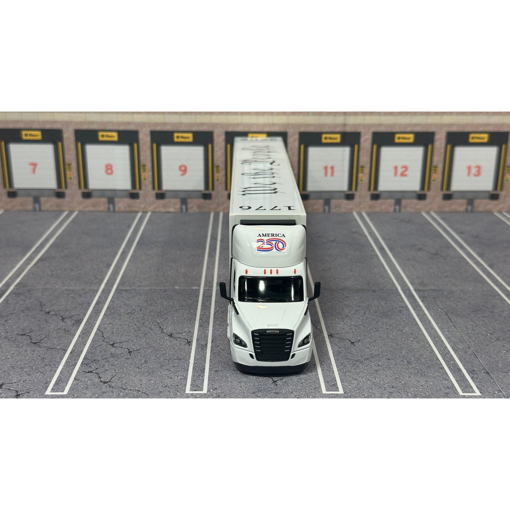 A diecast model of a white Freightliner Cascadia semi-truck with a long trailer decorated with American flags and a '250th Anniversary' logo, parked in a gray parking lot with a truck dock backdrop.