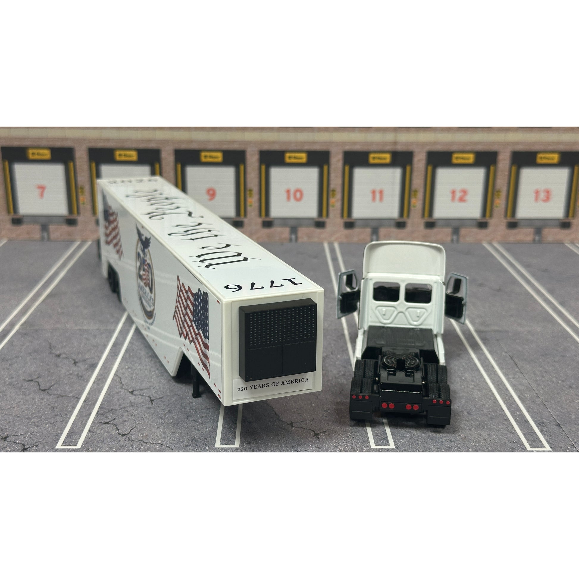 A diecast model of a white Freightliner Cascadia semi-truck with a long trailer decorated with American flags and a '250th Anniversary' logo, parked in a gray parking lot with a truck dock backdrop.
