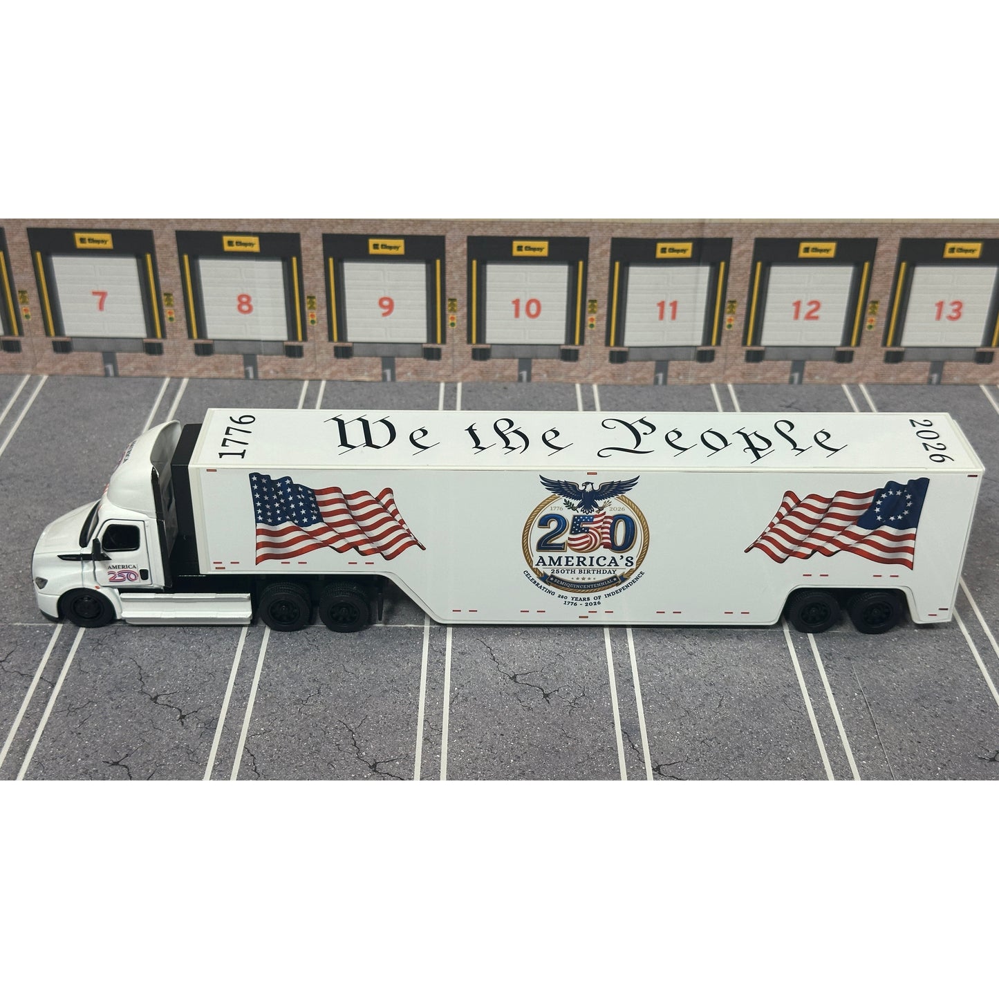 A diecast model of a white Freightliner Cascadia semi-truck with a long trailer decorated with American flags and a '250th Anniversary' logo, parked in a gray parking lot with a truck dock backdrop.