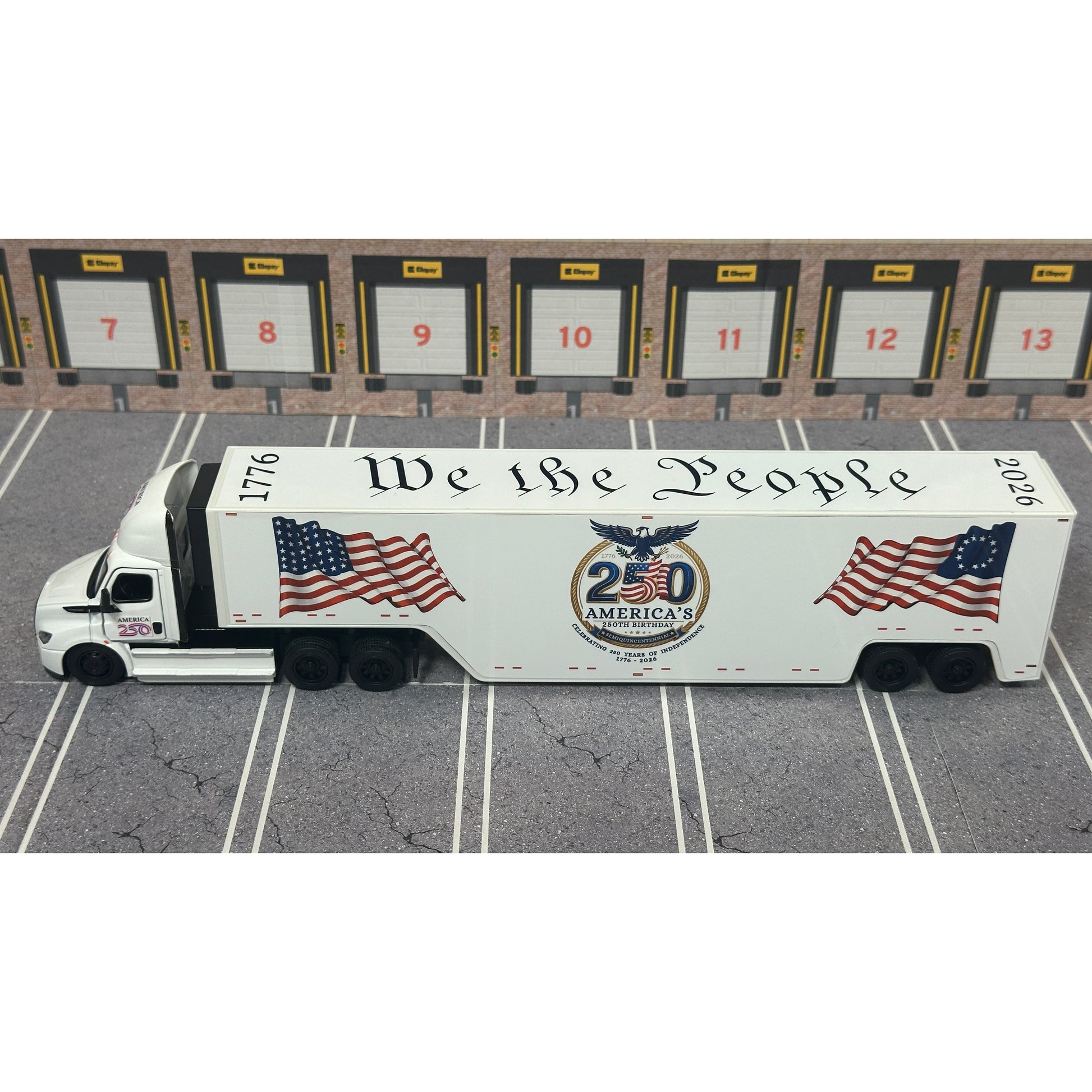 A diecast model of a white Freightliner Cascadia semi-truck with a long trailer decorated with American flags and a '250th Anniversary' logo, parked in a gray parking lot with a truck dock backdrop.