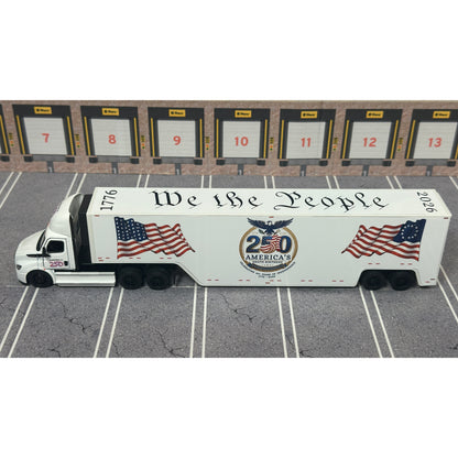 A diecast model of a white Freightliner Cascadia semi-truck with a long trailer decorated with American flags and a '250th Anniversary' logo, parked in a gray parking lot with a truck dock backdrop.