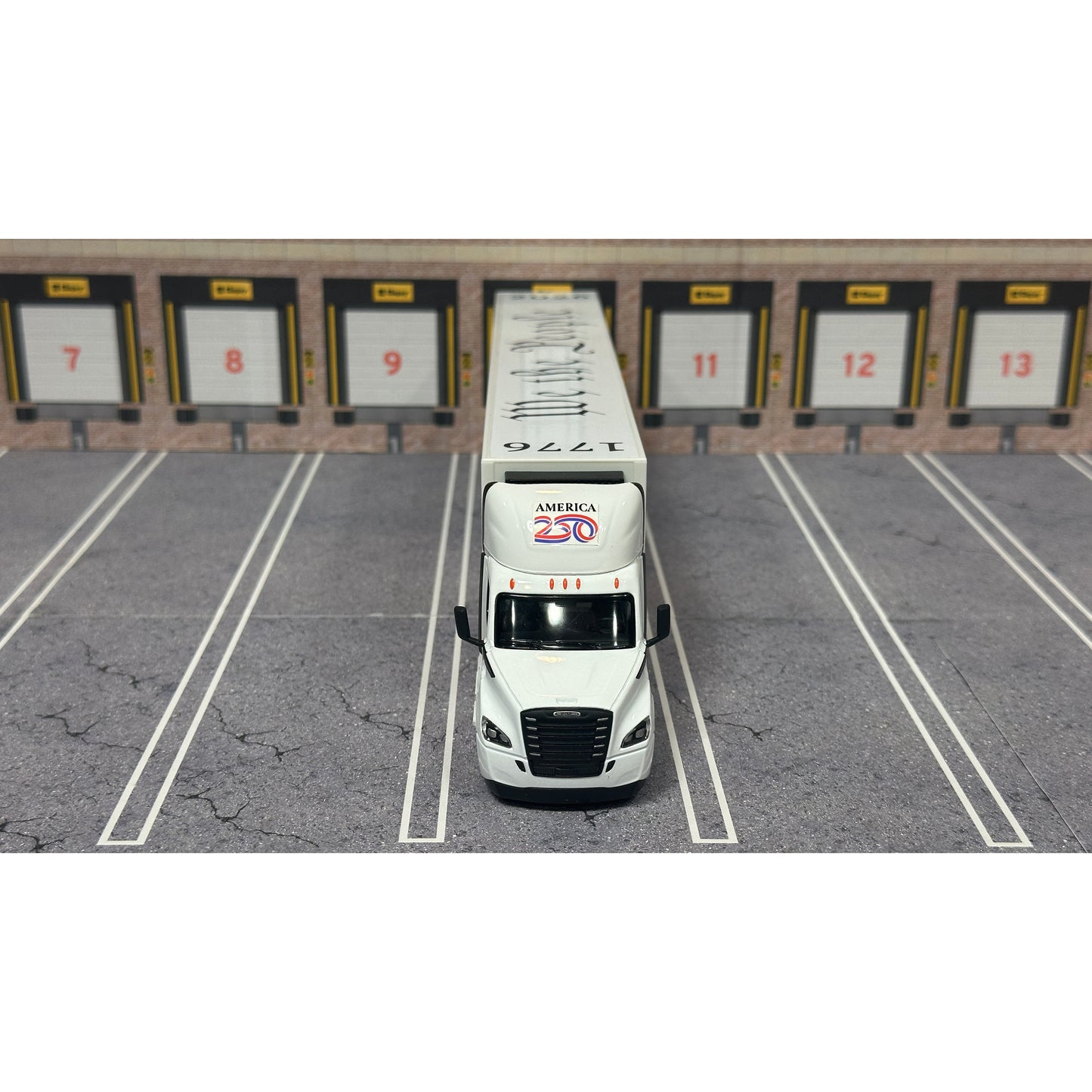 A diecast model of a Freightliner Cascadia semi-truck parked in a simulated parking lot. The trailer features a detailed historical design celebrating the United States' 250th anniversary with 'We the People' written on the top, alongside an American flag, eagle, and city skyline graphic with the text 'Land of the Free Home of the Brave' and 'United We Stand'.