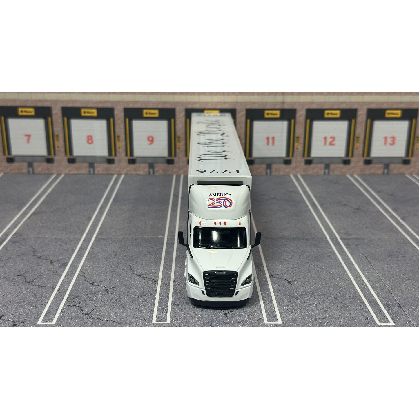 A diecast model of a Freightliner Cascadia semi-truck parked in a simulated parking lot. The trailer features a detailed historical design celebrating the United States' 250th anniversary with 'We the People' written on the top, alongside an American flag, eagle, and city skyline graphic with the text 'Land of the Free Home of the Brave' and 'United We Stand'.