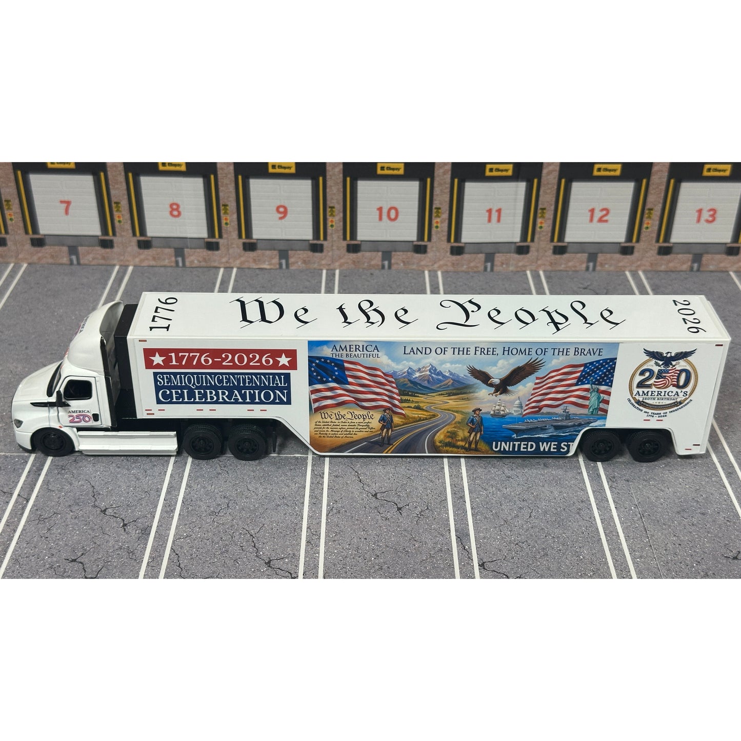 A diecast model of a Freightliner Cascadia semi-truck parked in a simulated parking lot. The trailer features a detailed historical design celebrating the United States' 250th anniversary with 'We the People' written on the top, alongside an American flag, eagle, and city skyline graphic with the text 'Land of the Free Home of the Brave' and 'United We Stand'.