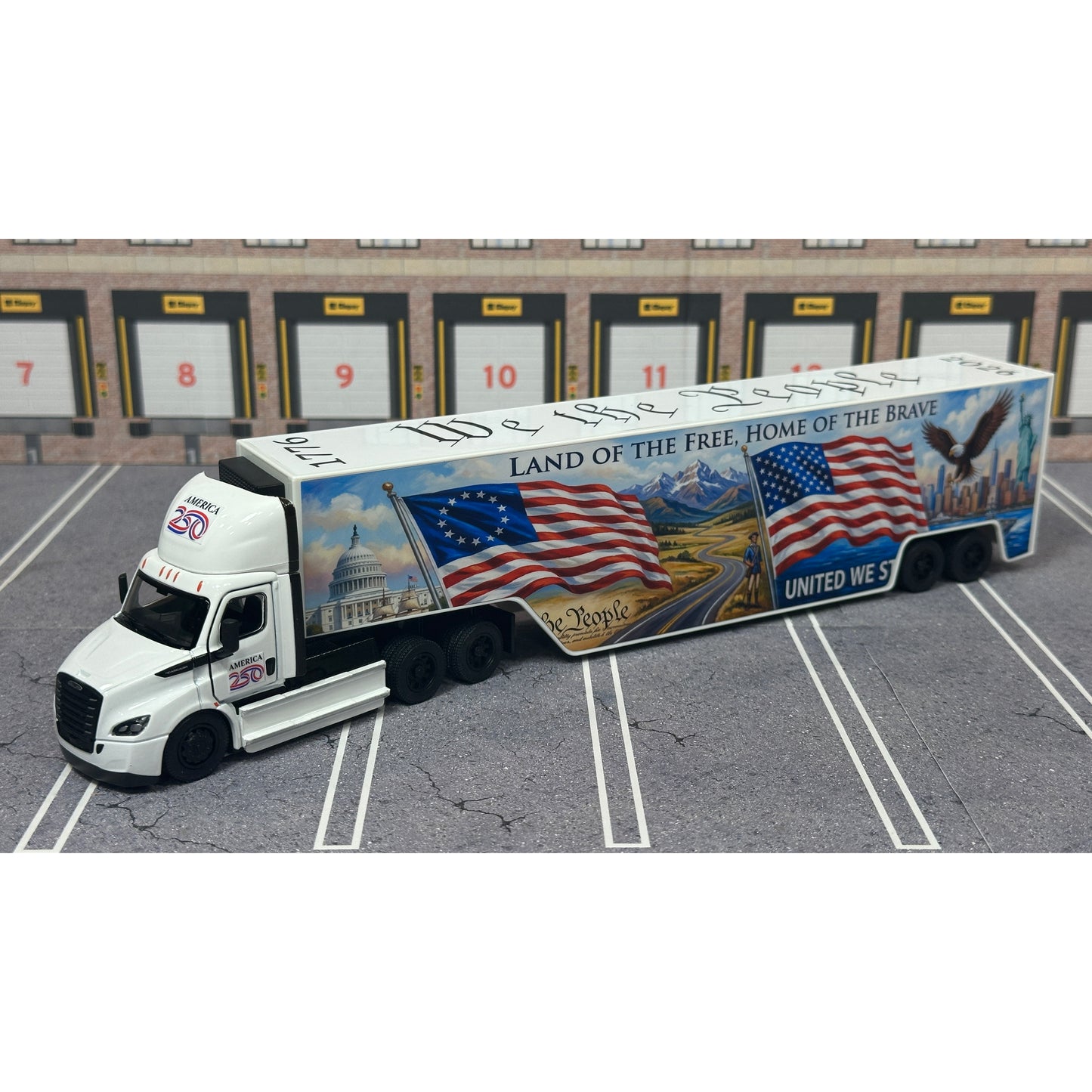 A diecast model of a Freightliner Cascadia semi-truck parked in a simulated parking lot. The trailer features a detailed historical design celebrating the United States' 250th anniversary with 'We the People' written on the top, alongside an American flag, eagle, and city skyline graphic with the text 'Land of the Free Home of the Brave' and 'United We Stand'.