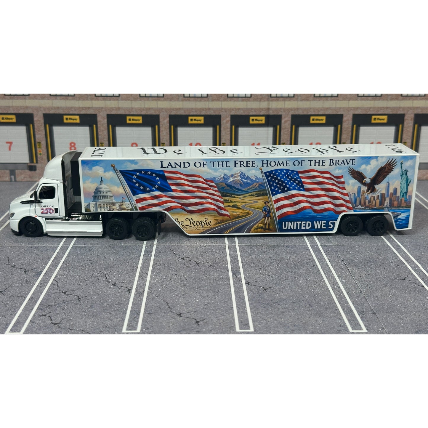 A diecast model of a Freightliner Cascadia semi-truck parked in a simulated parking lot. The trailer features a detailed historical design celebrating the United States' 250th anniversary with 'We the People' written on the top, alongside an American flag, eagle, and city skyline graphic with the text 'Land of the Free Home of the Brave' and 'United We Stand'.