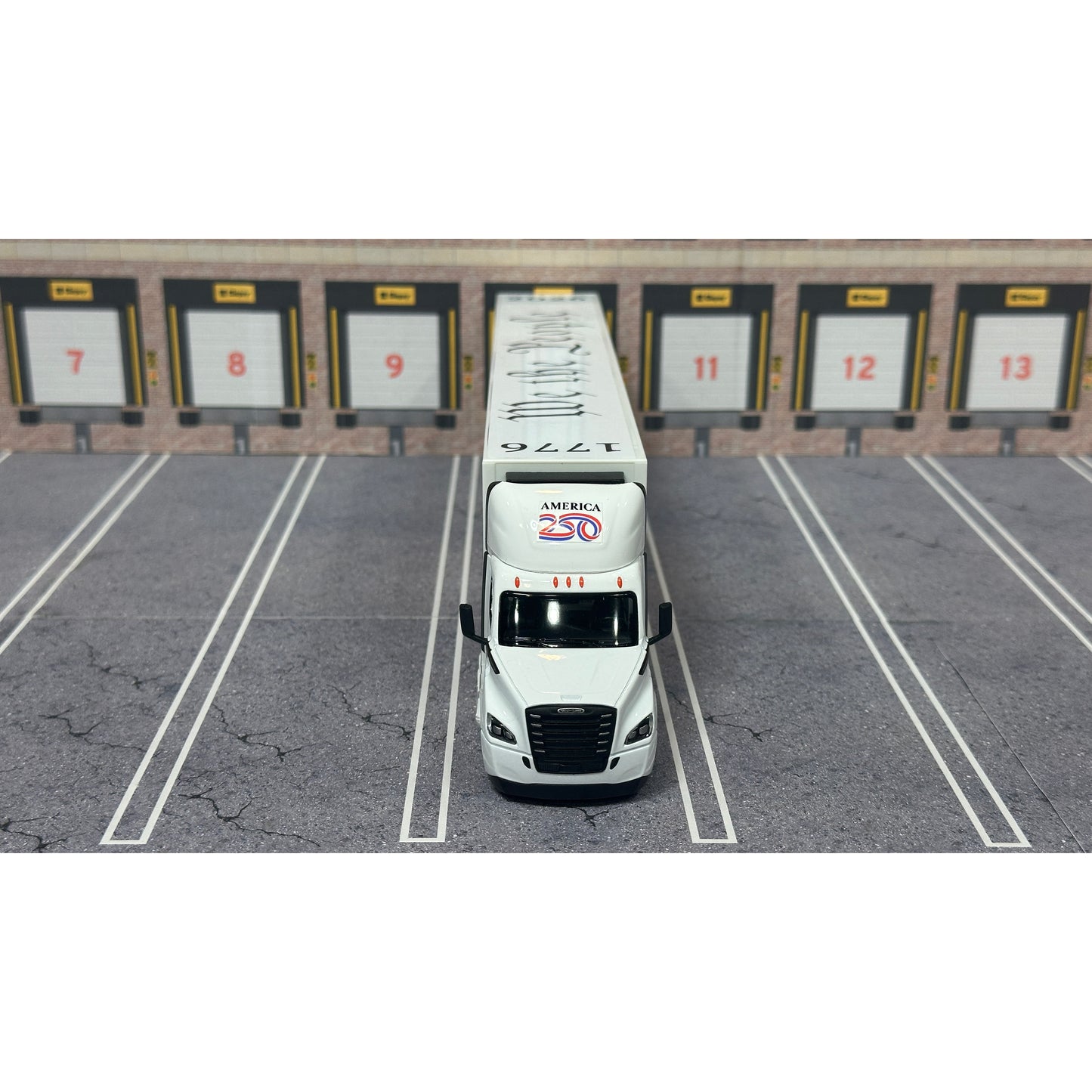 A diecast model of a Freightliner Cascadia semi-truck parked in a simulated parking lot. The trailer features a detailed historical design celebrating the United States' 250th anniversary with 'We the People' written on the top, alongside an American flag, eagle, and city skyline graphic with the text 'Land of the Free Home of the Brave' and 'United We Stand'.