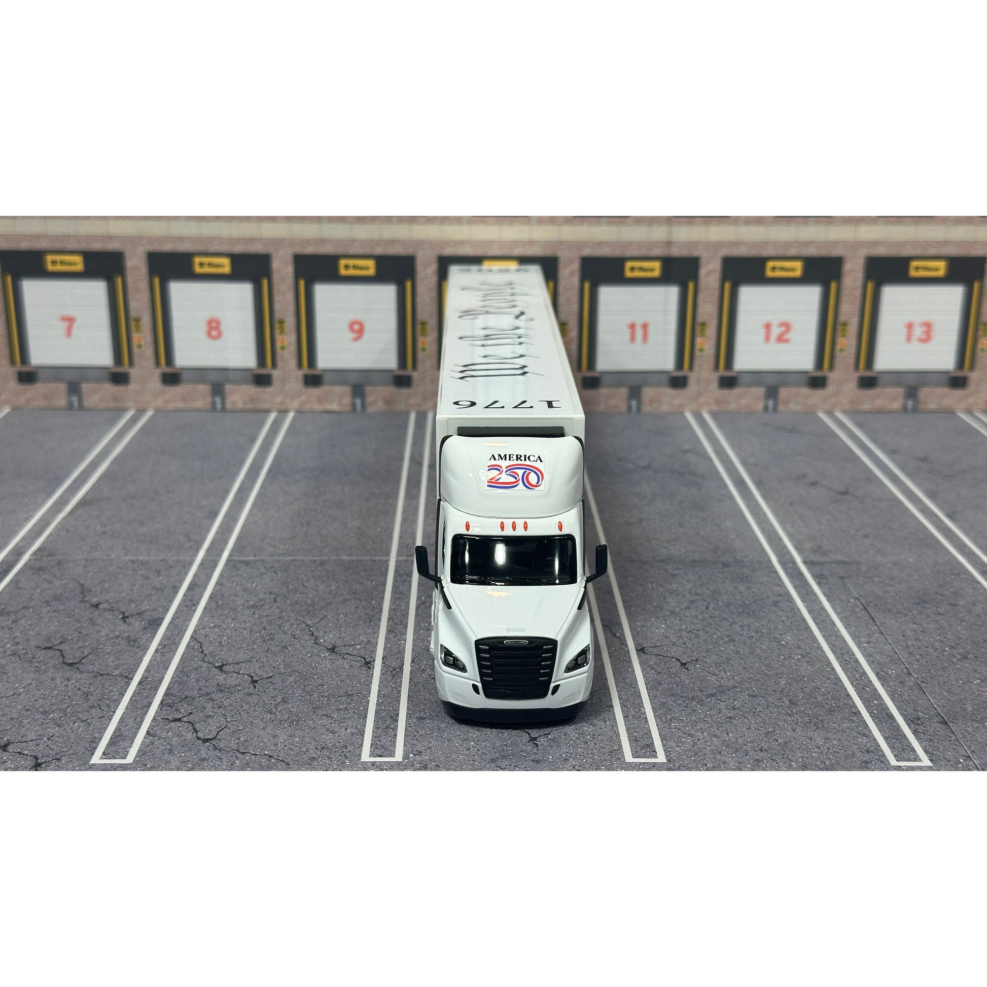 A diecast model of a Freightliner Cascadia semi-truck parked in a simulated parking lot. The trailer features a detailed historical design celebrating the United States' 250th anniversary with 'We the People' written on the top, alongside an American flag, eagle, and city skyline graphic with the text 'Land of the Free Home of the Brave' and 'United We Stand'.