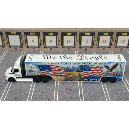 A diecast model of a Freightliner Cascadia semi-truck parked in a simulated parking lot. The trailer features a detailed historical design celebrating the United States' 250th anniversary with 'We the People' written on the top, alongside an American flag, eagle, and city skyline graphic with the text 'Land of the Free Home of the Brave' and 'United We Stand'.