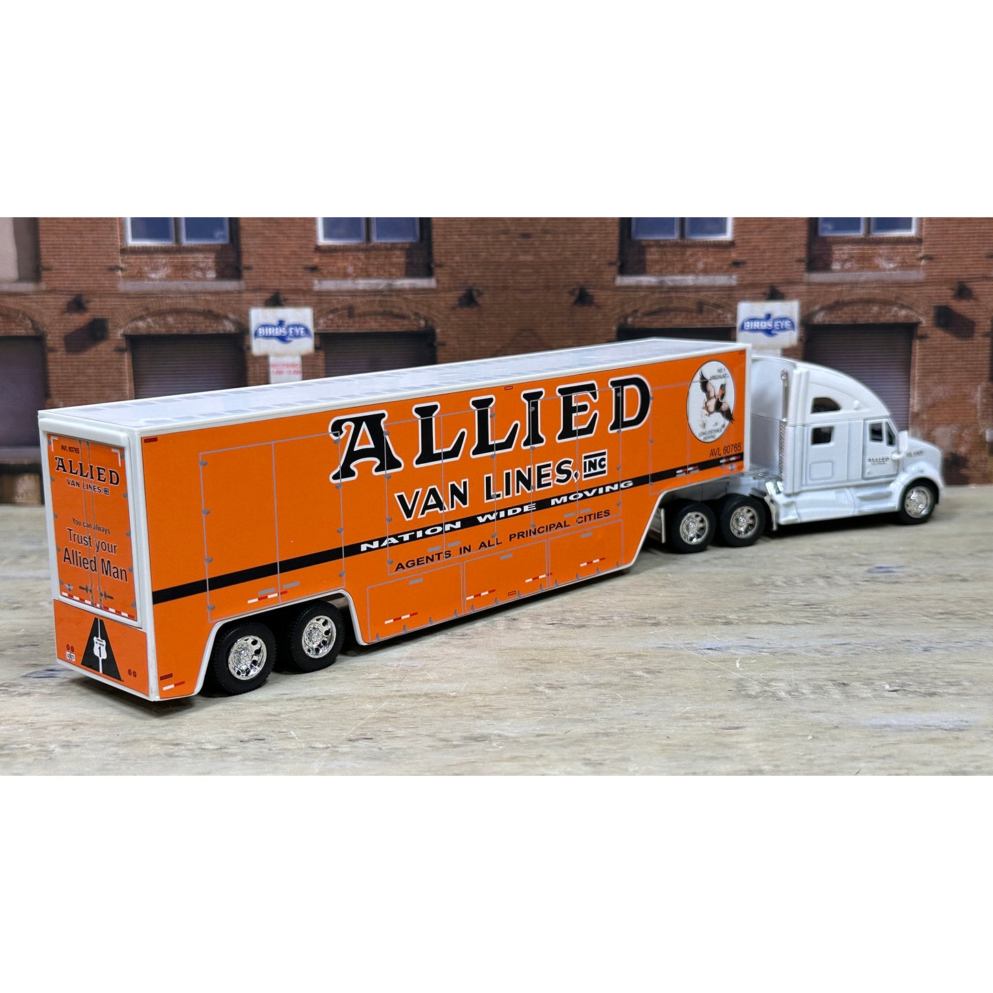 Model of a semi-truck with an orange trailer labeled 'Allied Van Lines' in front of a brick building.