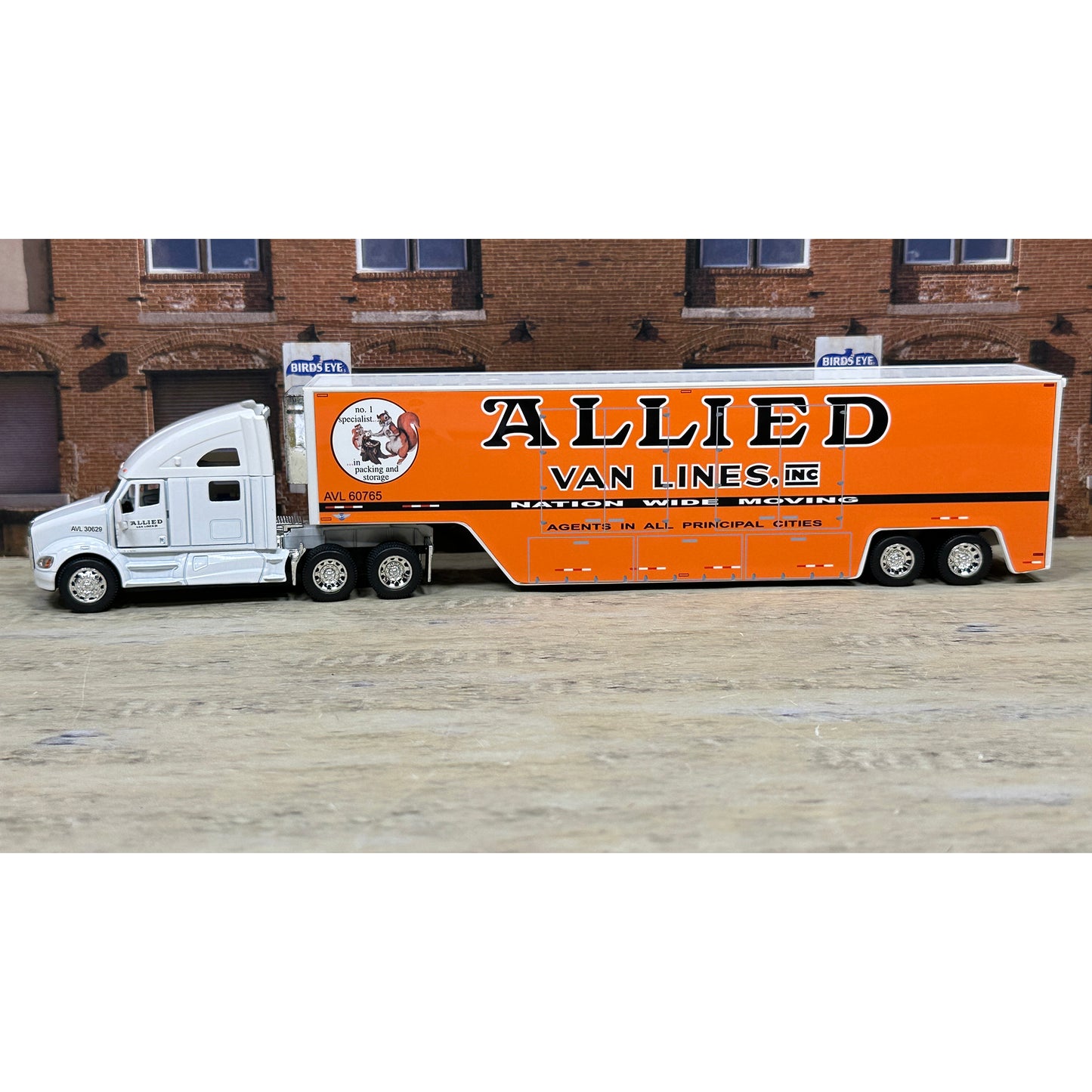 Model of a semi-truck with an orange trailer labeled 'Allied Van Lines' in front of a brick building.