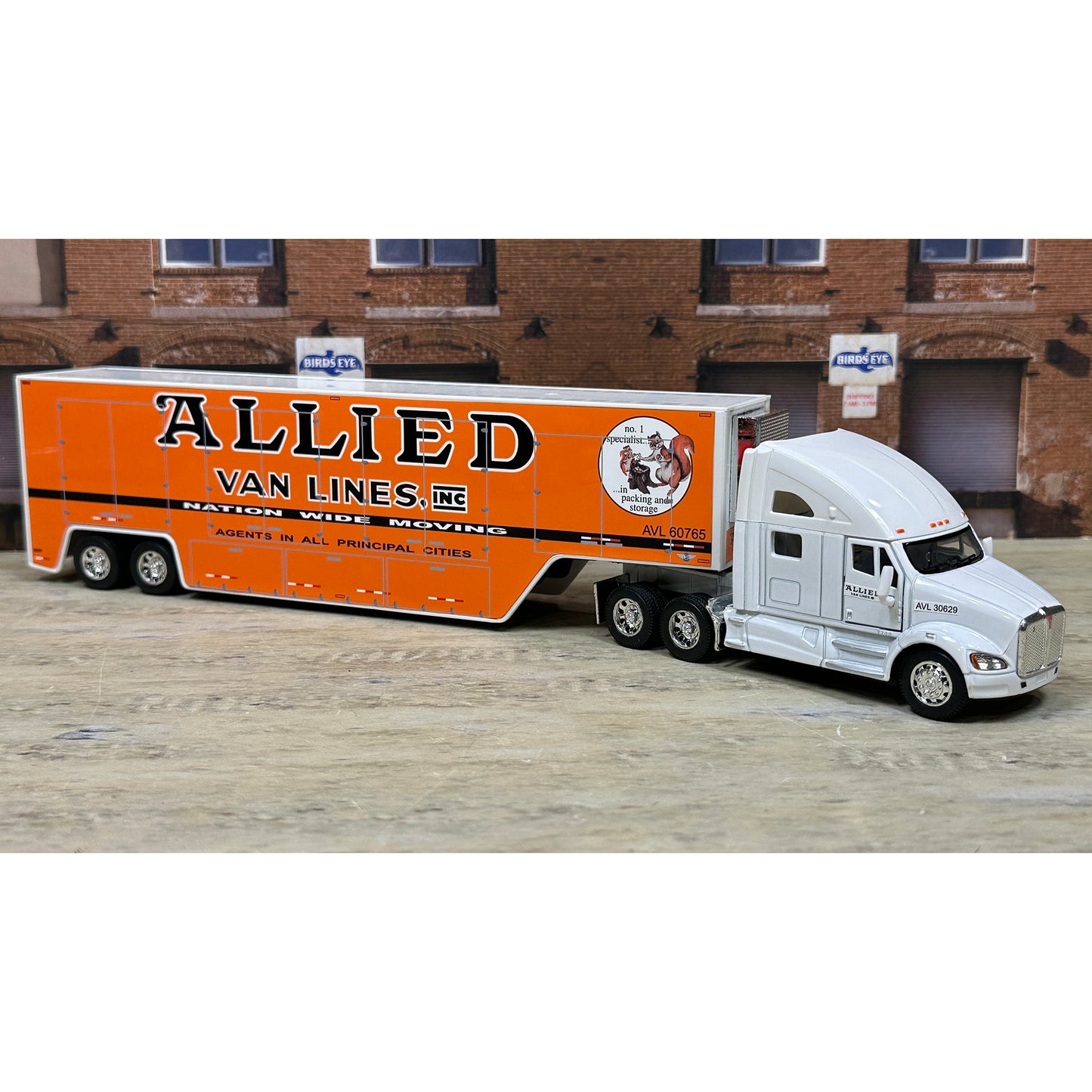 Model of a semi-truck with an orange trailer labeled 'Allied Van Lines' in front of a brick building.