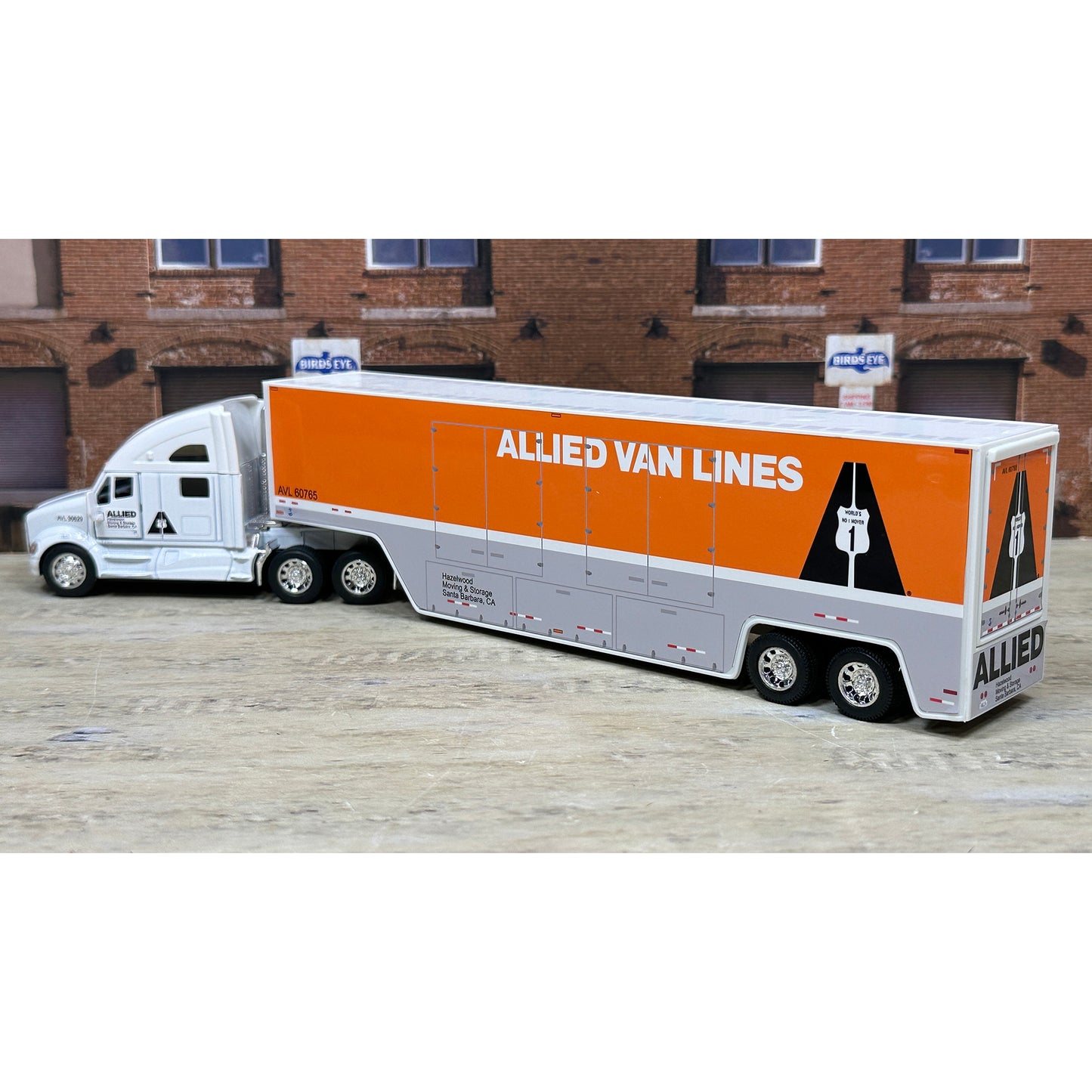 Model of a semi-truck with an orange trailer labeled 'Allied Van Lines' in front of a brick building.